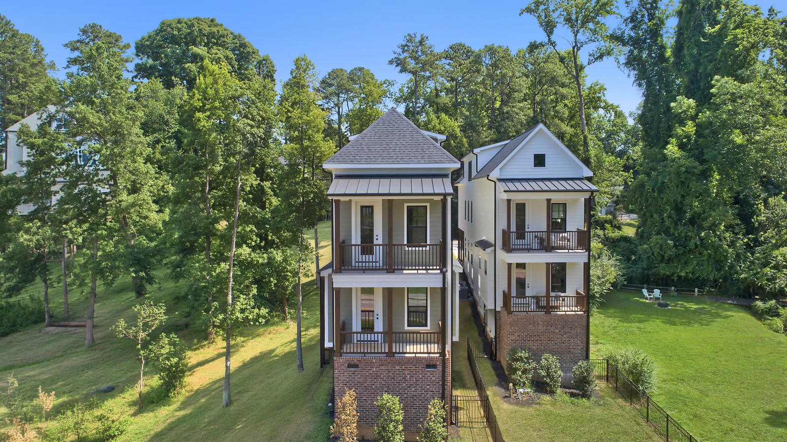 Two tall, narrow new homes with stacked front porches and brick bases among mature trees