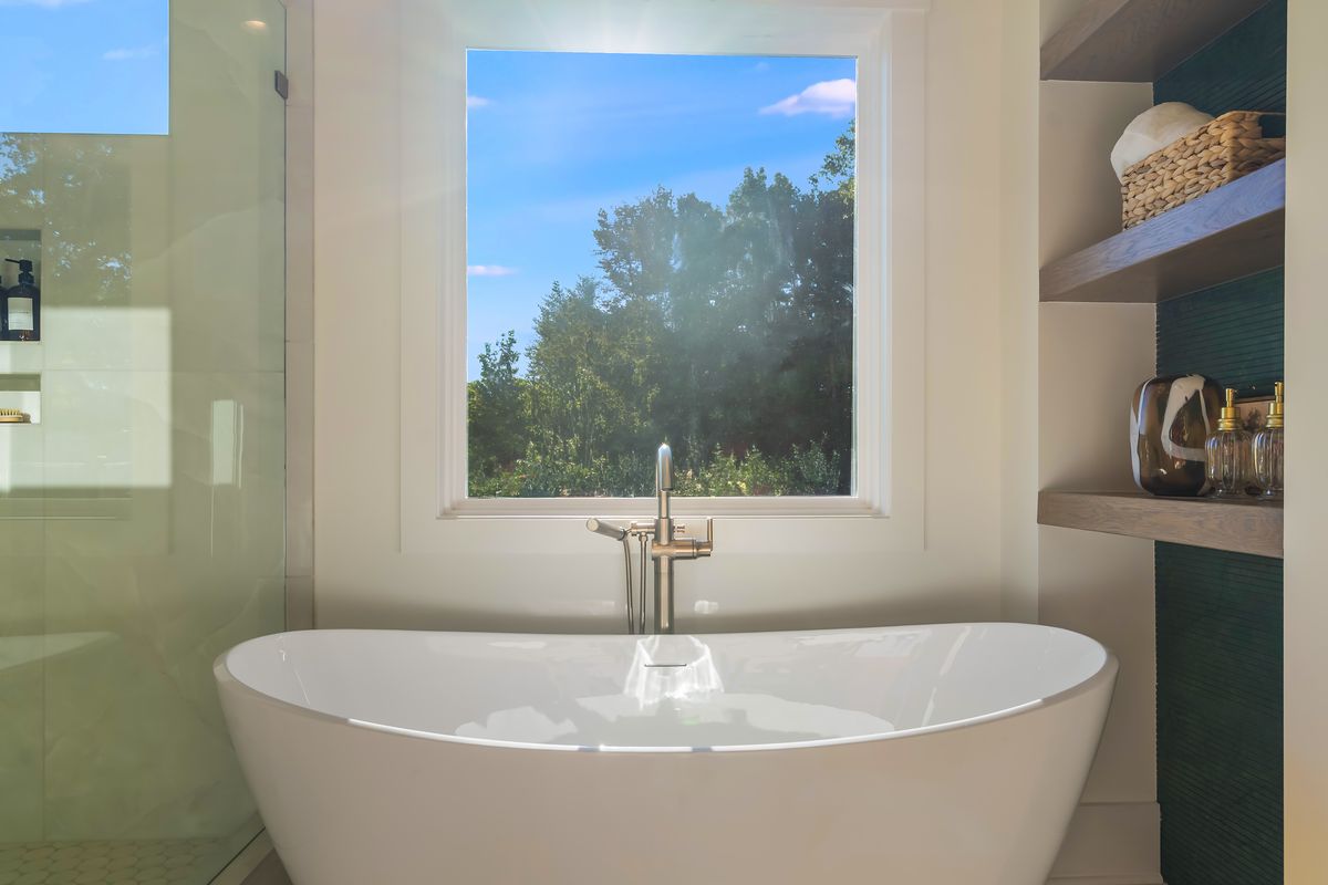 A freestanding tub with a floor-mounted filler beneath a window, beside open shelving