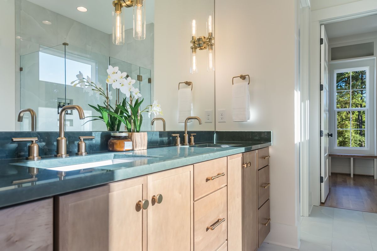 A double vanity with light wood cabinets, a dark green countertop, brass faucets and sconces, and an orchid