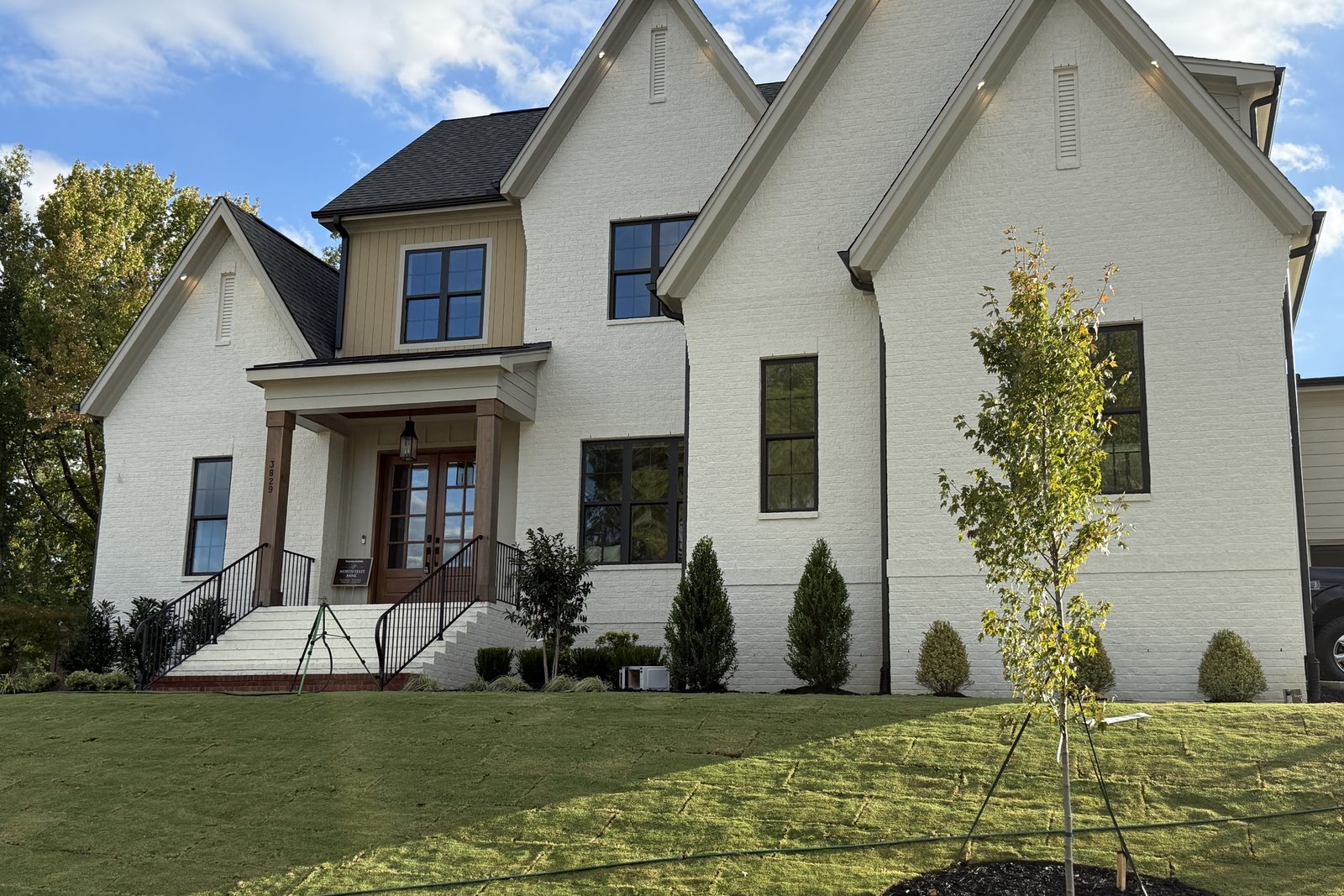 A painted white brick home with three steep gables and a cedar-post porch, on a new lawn